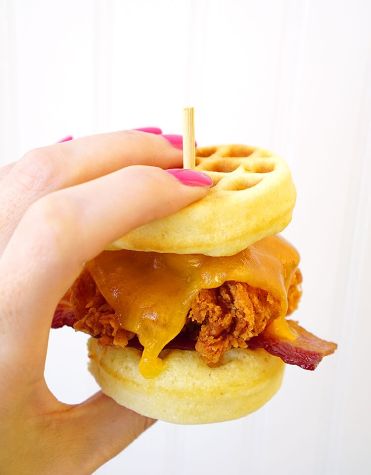 hand holding a chicken waffle slider sandwich with melted cheese and bacon