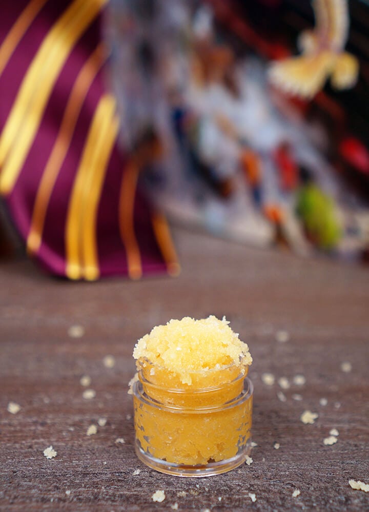 Small jar of Butterbeer sugar scrub on wood background with Harry Potter book and Gryffindor tie in background