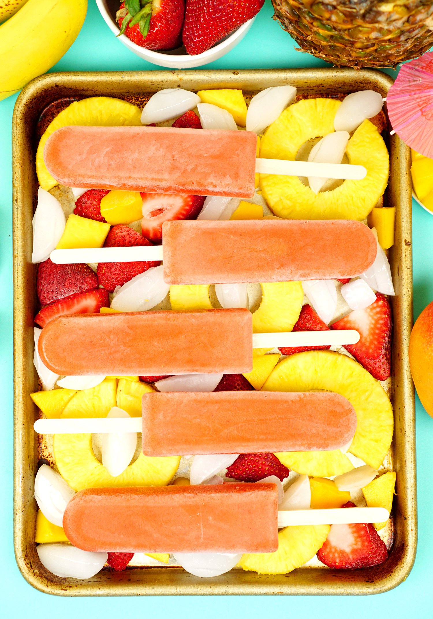 Tropical smoothie popsicles on a tray of ice with sliced pineapple rings and strawberries