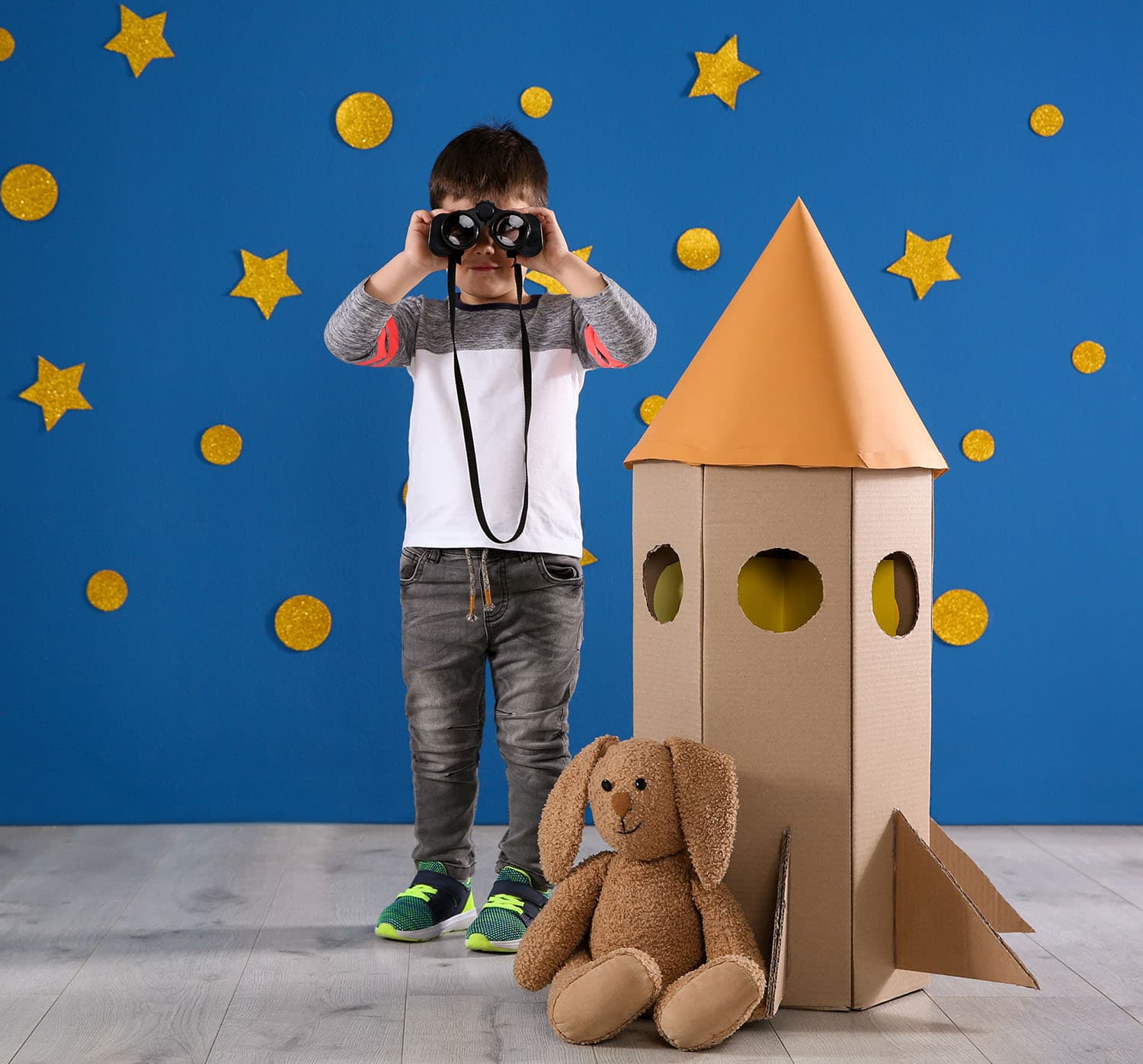 child holding binoculars to face with cardboard rocket ship and stuffed animal
