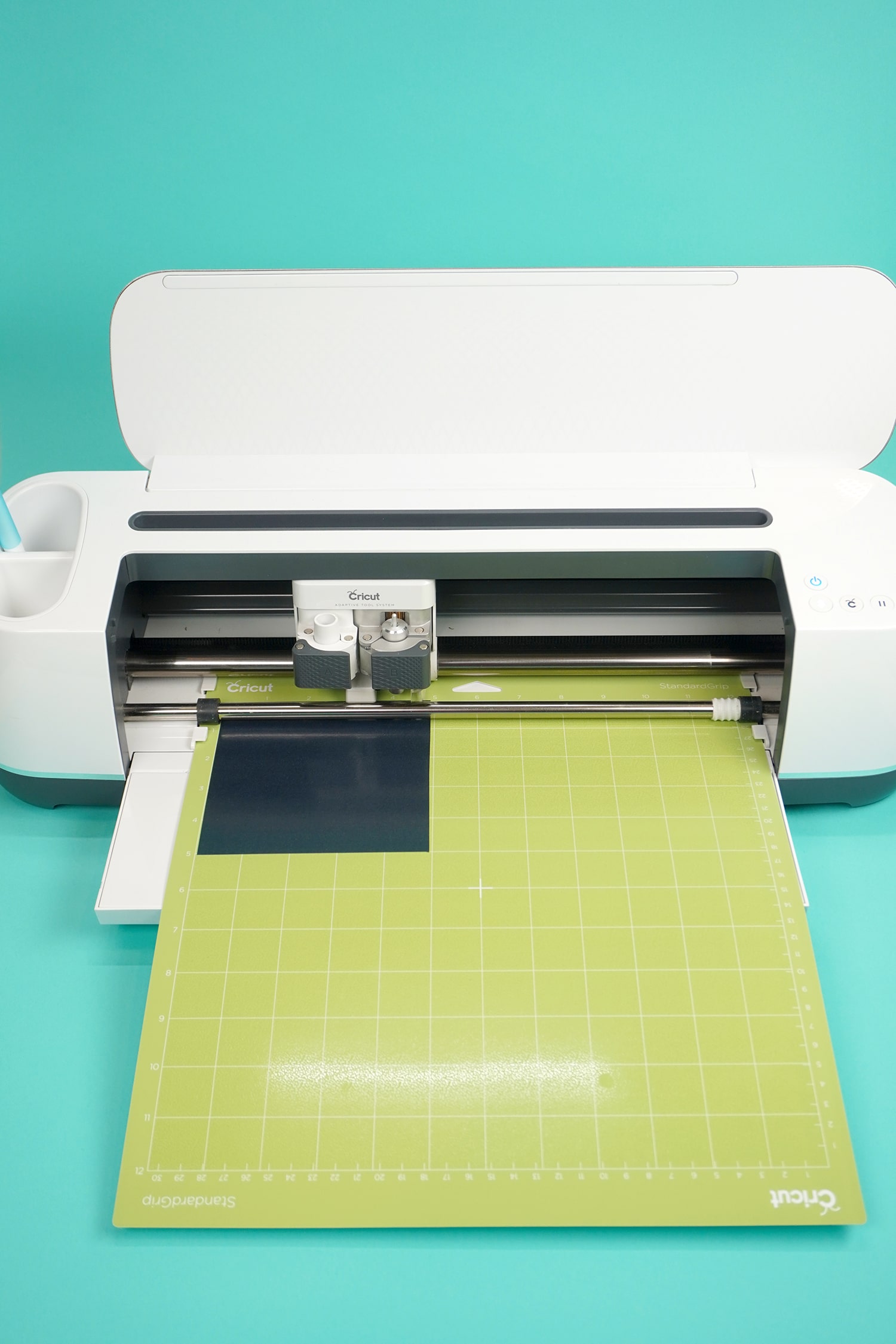A close up of a white Cricut maker with a green cutting mat on a turquoise background 