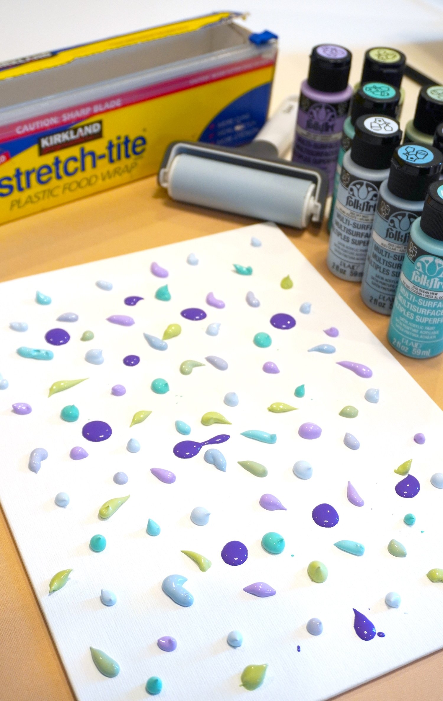 Paint drops on canvas with plastic wrap, brayer, and bottles of paint in background