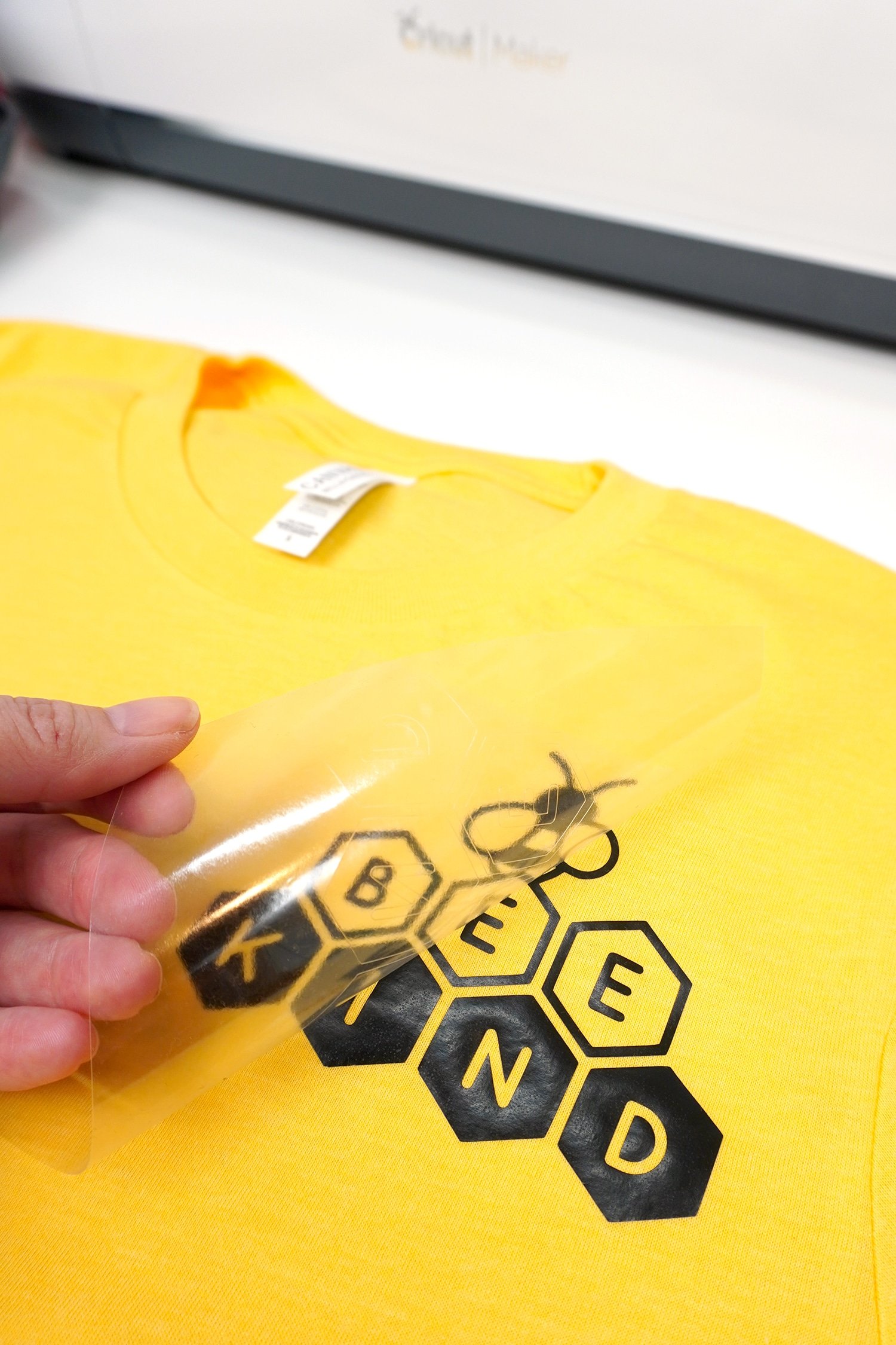 pulling off iron on backing plastic to reveal Bee Kind image on yellow tshirt