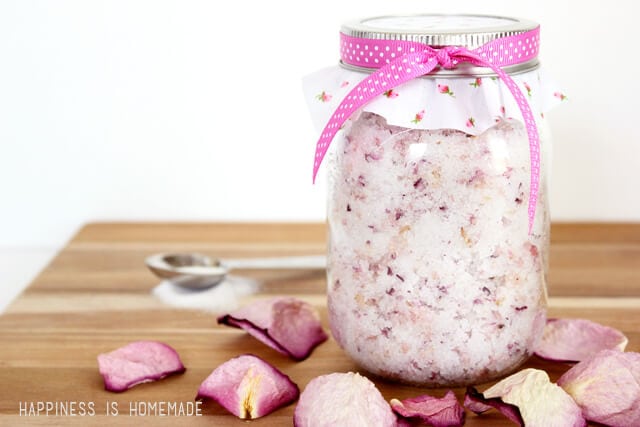 Glass jar of rose petal sugar scrub on a wood background surrounded by rose petals, a spoon, and a small pile of sugar