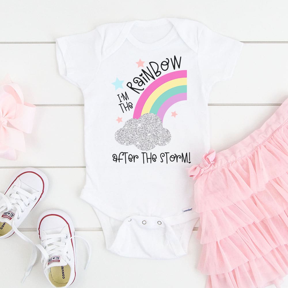 \"I\'m the Rainbow After the Storm\" SVG on a white baby onesie on wood background with pink tutu and white baby shoes
