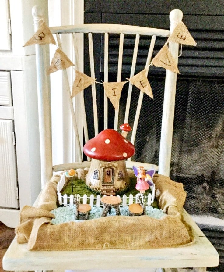 Red mushroom shaped fairy garden house with fairy figurine in burlap lined box sitting on a white wood chair 