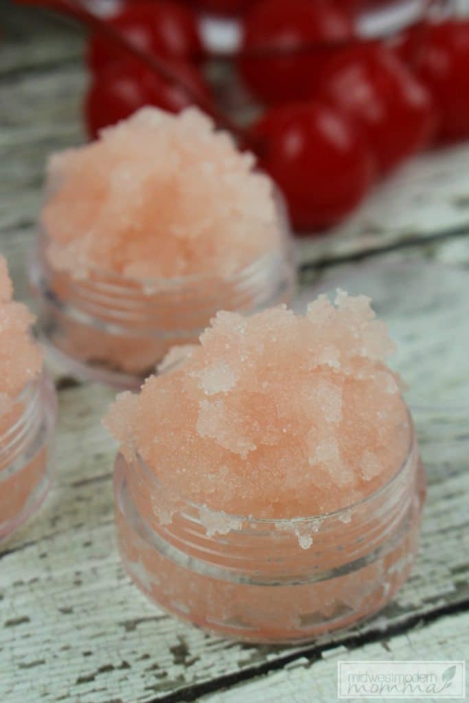 Close up of a small jar of pink cherry vanilla lip scrub with cherries in the background