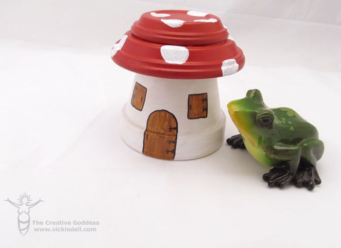 flower pot shaped fairy garden house painted to look like mushroom with red and white polka dot top with a green ceramic frog next to it