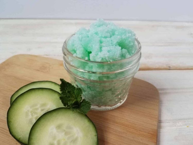 A jar of green cucumber mint sugar scrub on a wood background with cucumber slices and mint leaves