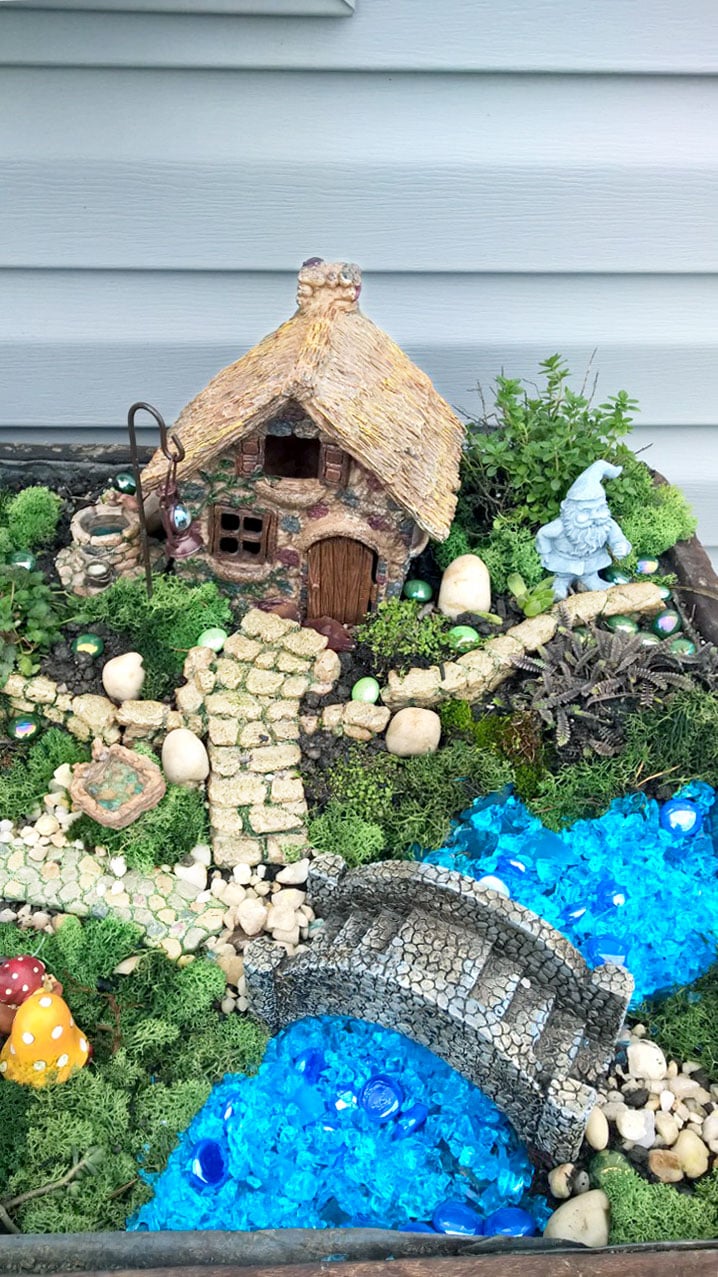 Fairy garden cottage in planter with greenery, rock bridge, and blue stones to simulate water