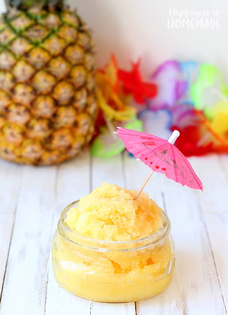 Yellow pina colada pineapple sugar scrub in a glass jar with a pink cocktail umbrella on top and a pineapple and Hawaiian lei in background