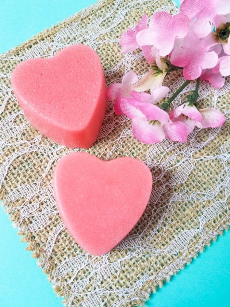 two pink heart shaped strawberry sugar scrub bars on an aqua and burlap background with pink flowers