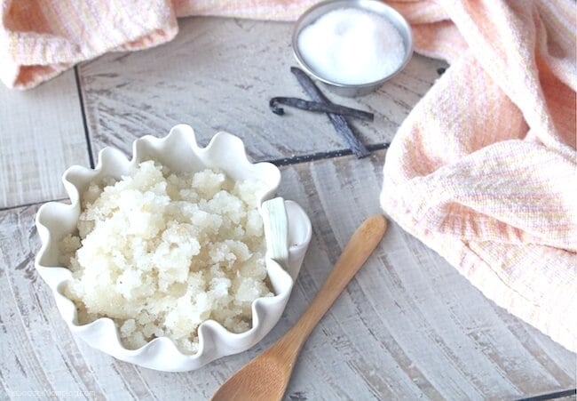 Bowl of vanilla honey lip scrub in wavy bowl on a wood background with small bowl of sugar, vanilla beans, and wooden spoon