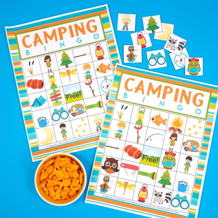 Two Camping Bingo Printable game cards on a blue background with scattered calling cards and bowl of Goldfish crackers for game markers