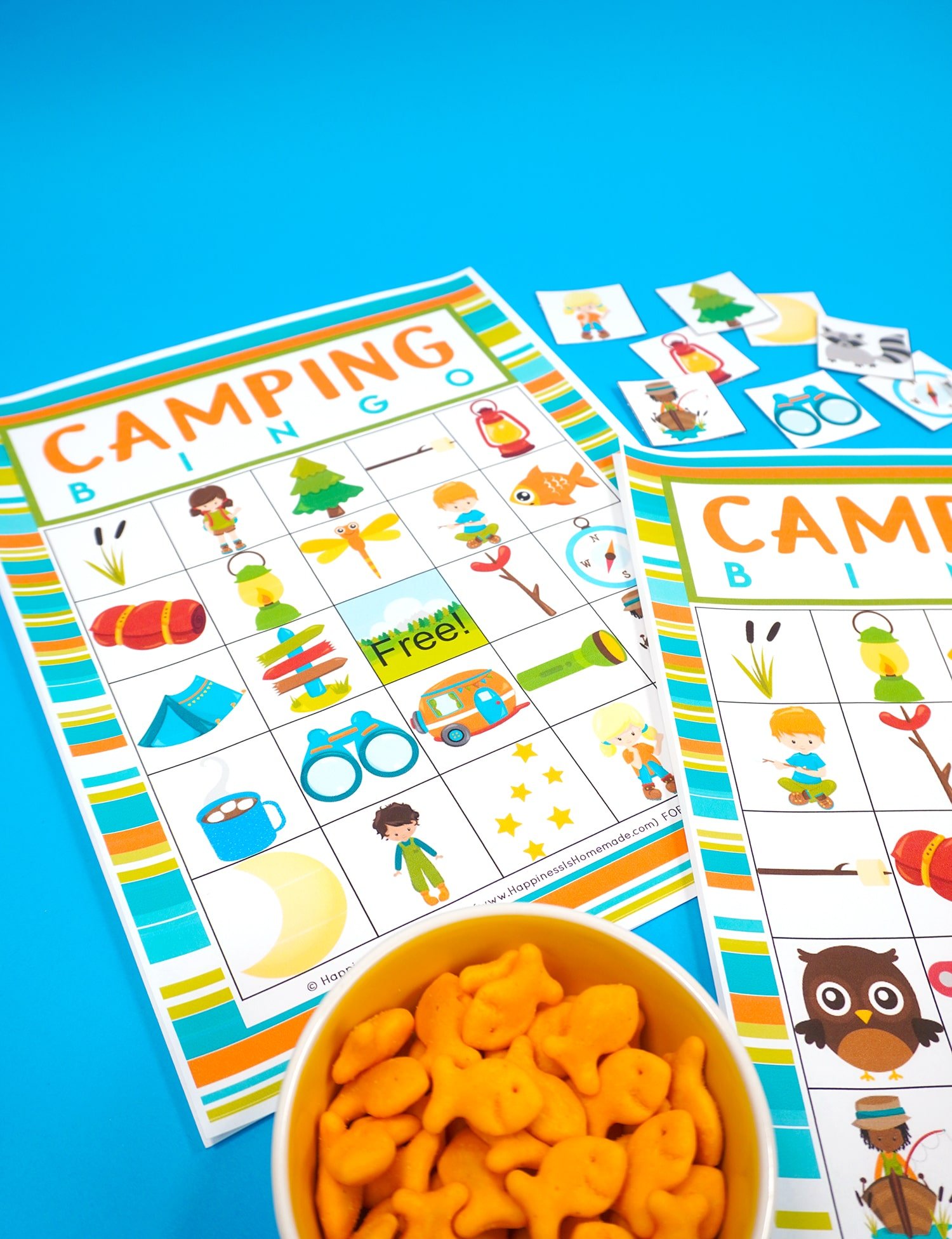 Two Camping Bingo Printable game cards on a blue background with scattered calling cards and bowl of Goldfish crackers for game markers