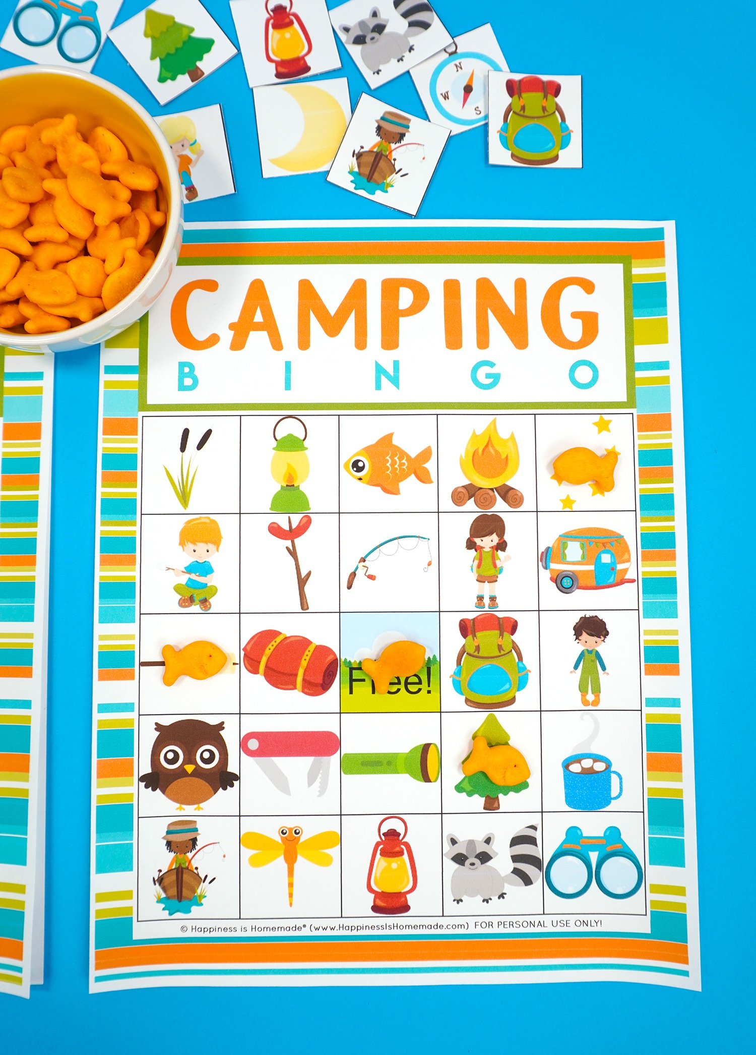 Camping Bingo Printable game card on a blue background with scattered calling cards and bowl of Goldfish crackers for game markers