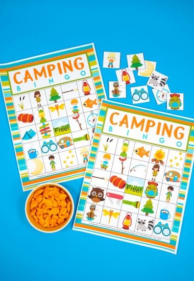 Two Camping Bingo Printable game cards on a blue background with scattered calling cards and bowl of Goldfish crackers for game markers