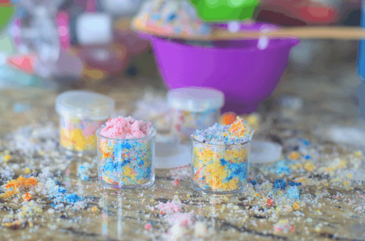 Two small jars of multicolored \"unicorn\" lip scrub with spilled sugar scrub all around in background