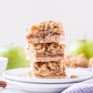 Three Apple Pie Bars stacked on a white plate with two green apples in the background