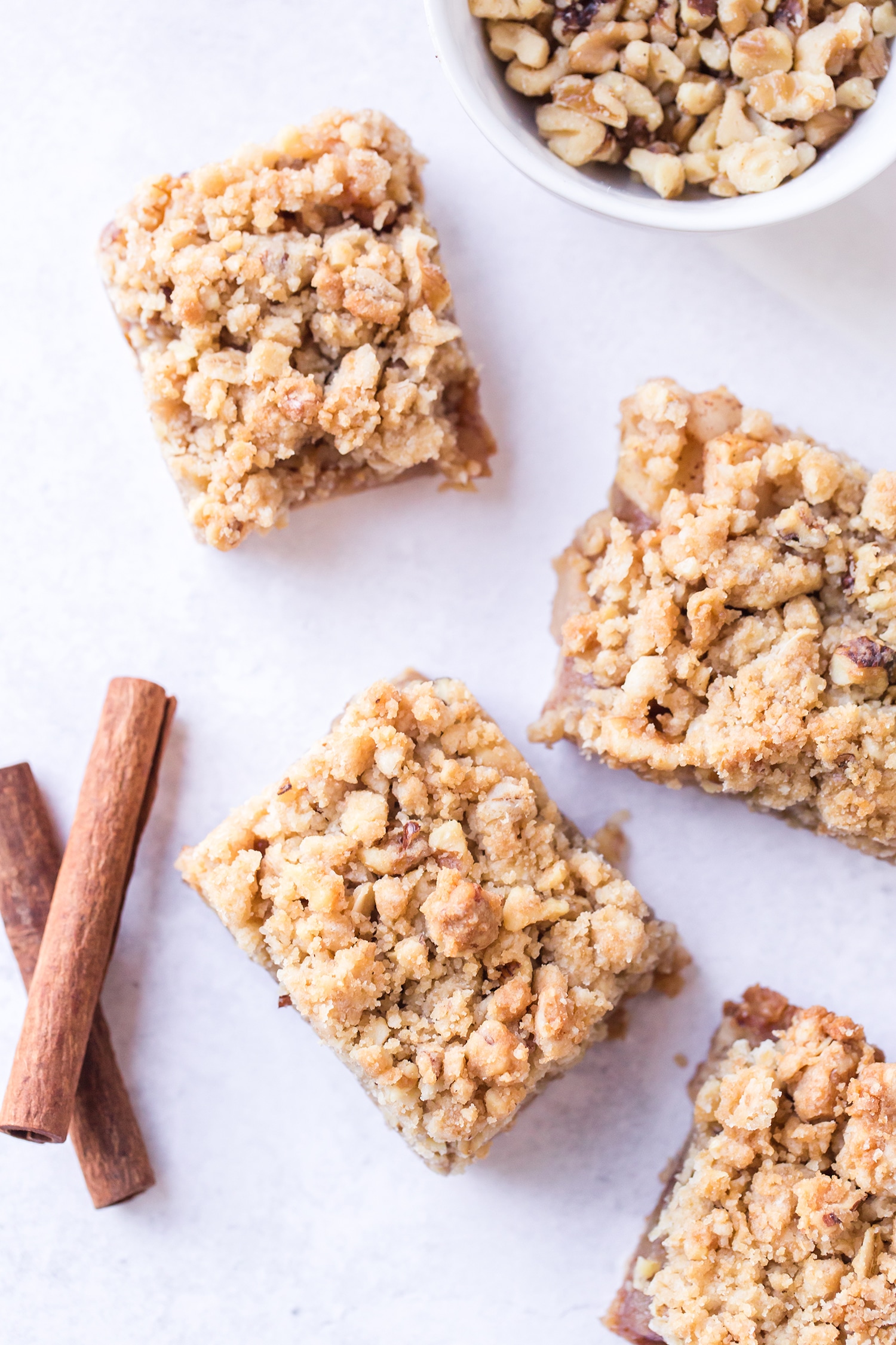 Four sliced apple pie bars on a white marble background with cinnamon sticks