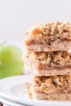 Three Apple Pie Bars stacked on a white plate with green apples in the background