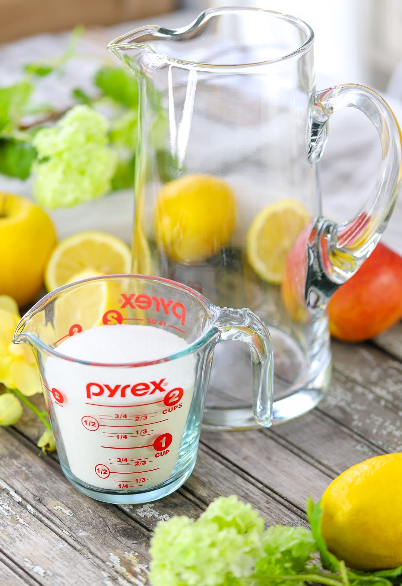 empty clear glass pitcher, measuring cup of sugar surrounded by apples and lemons