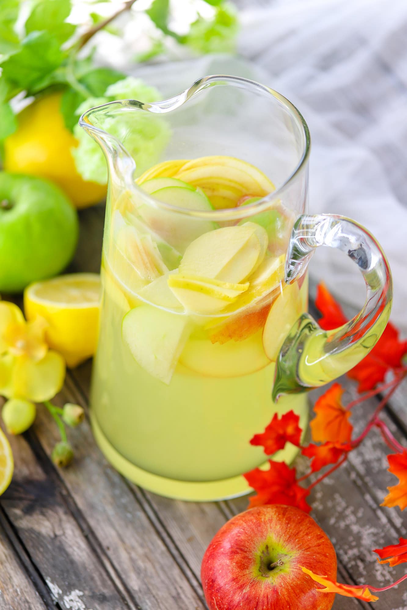 clear glass pitcher full of lemonade and apple slices surrounded by apples and lemons