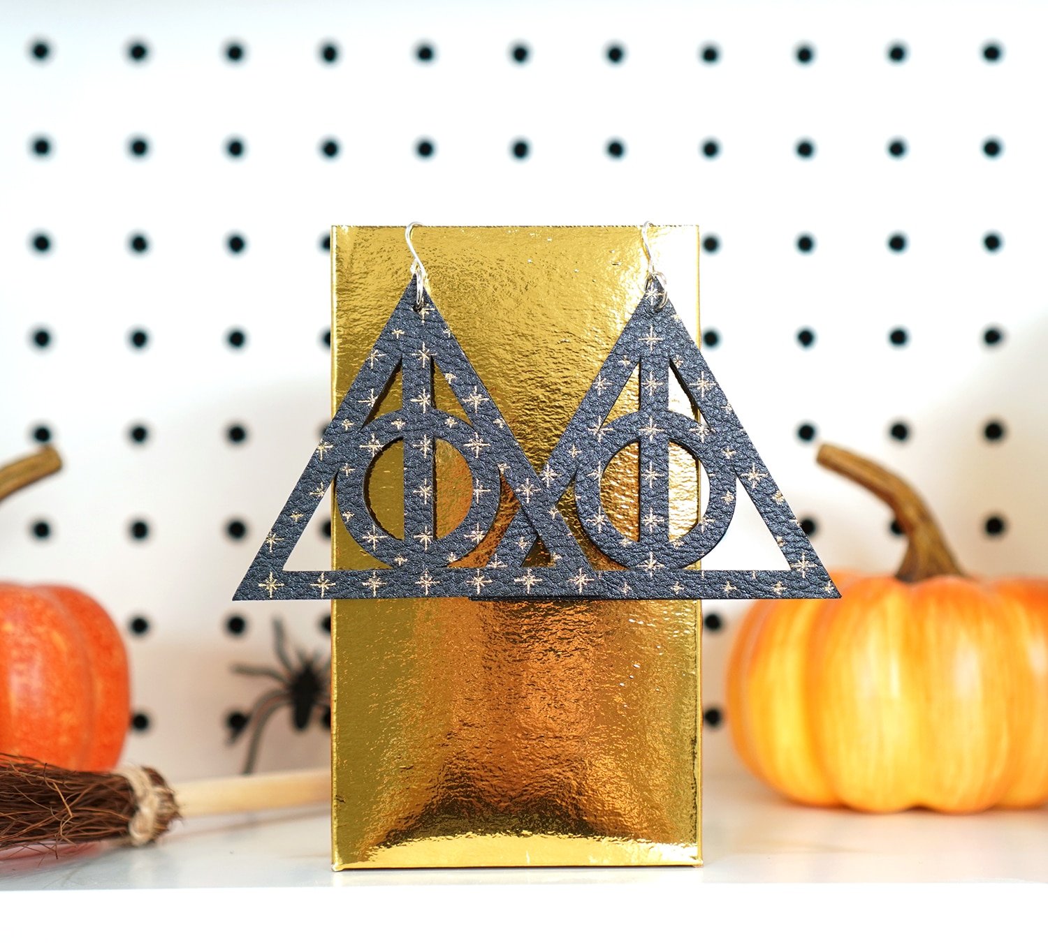 Deathly Hallows leather earrings with gold star pattern hanging on gold box with pumpkin in background