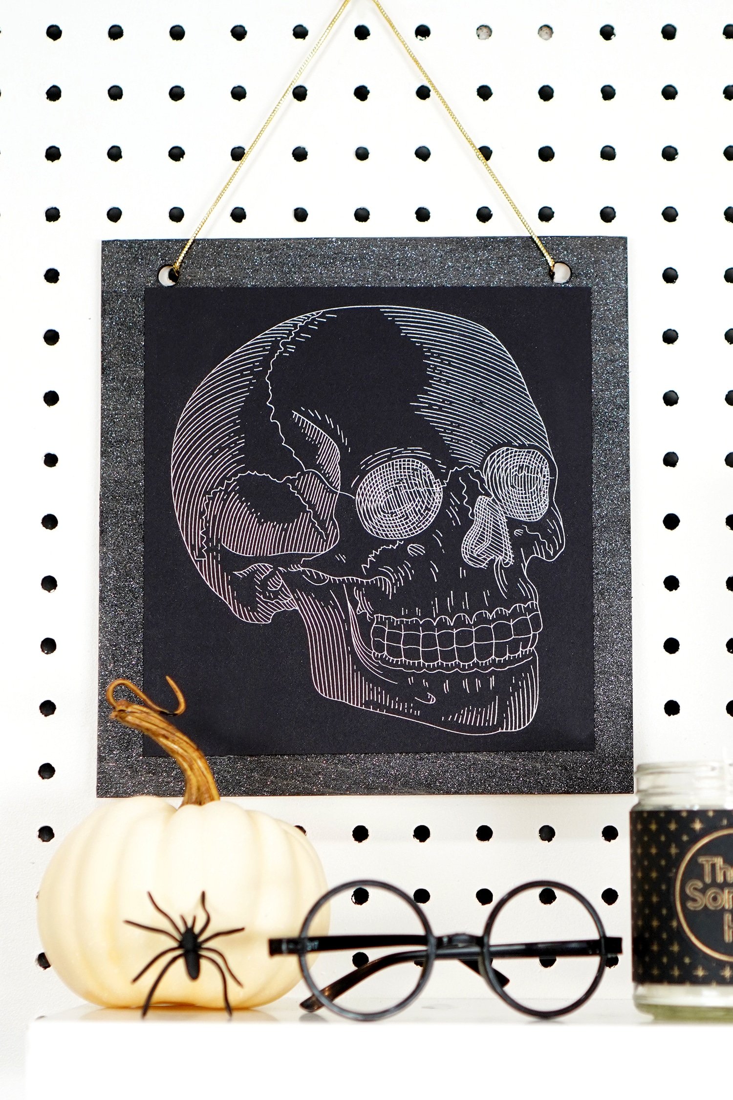 Silver foil skull art print hanging on pegboard with pumpkin, spider, and Harry Potter glasses in foreground