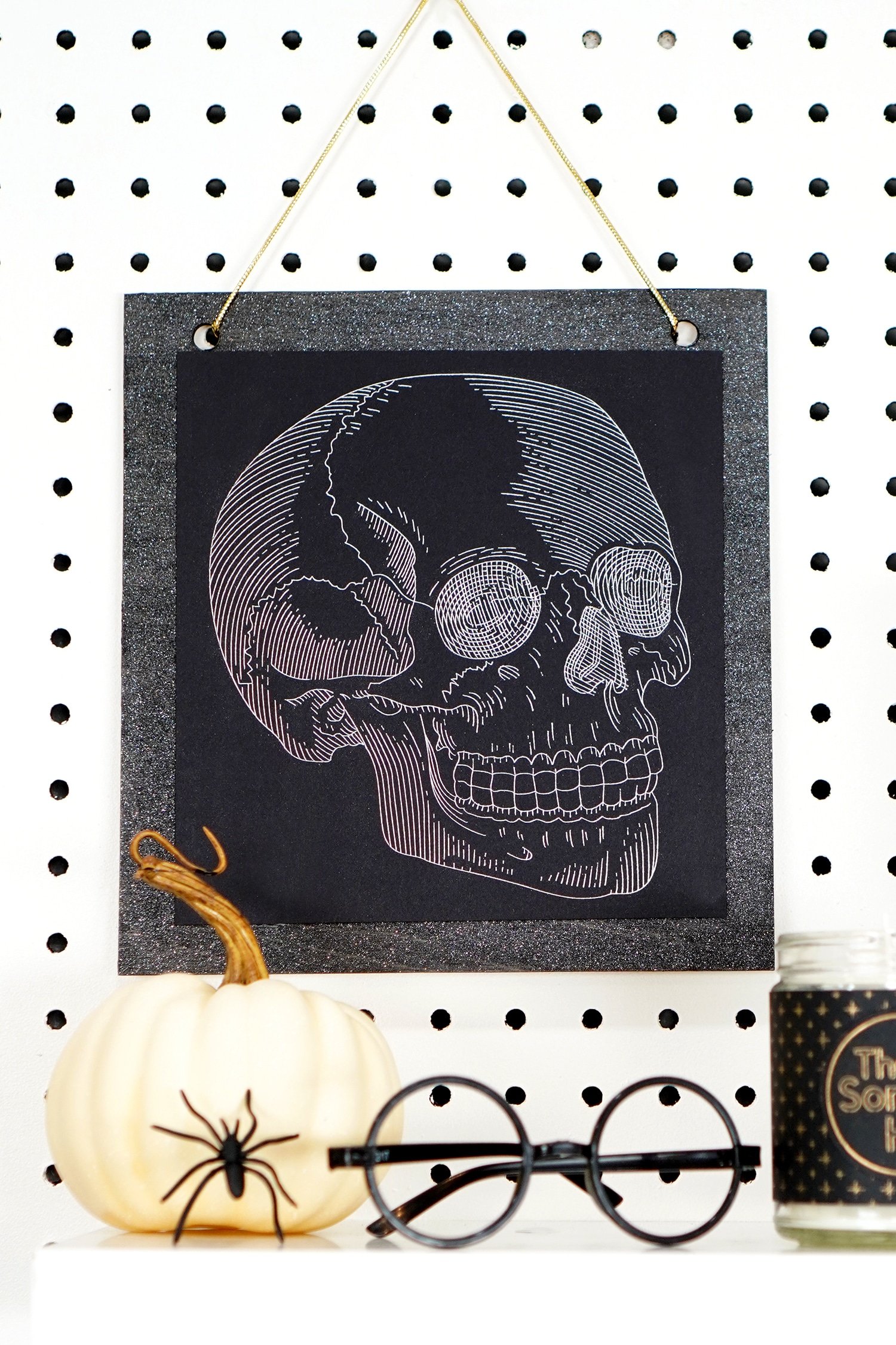 Silver foil skull art print hanging on pegboard with pumpkin, spider, and Harry Potter glasses in foreground