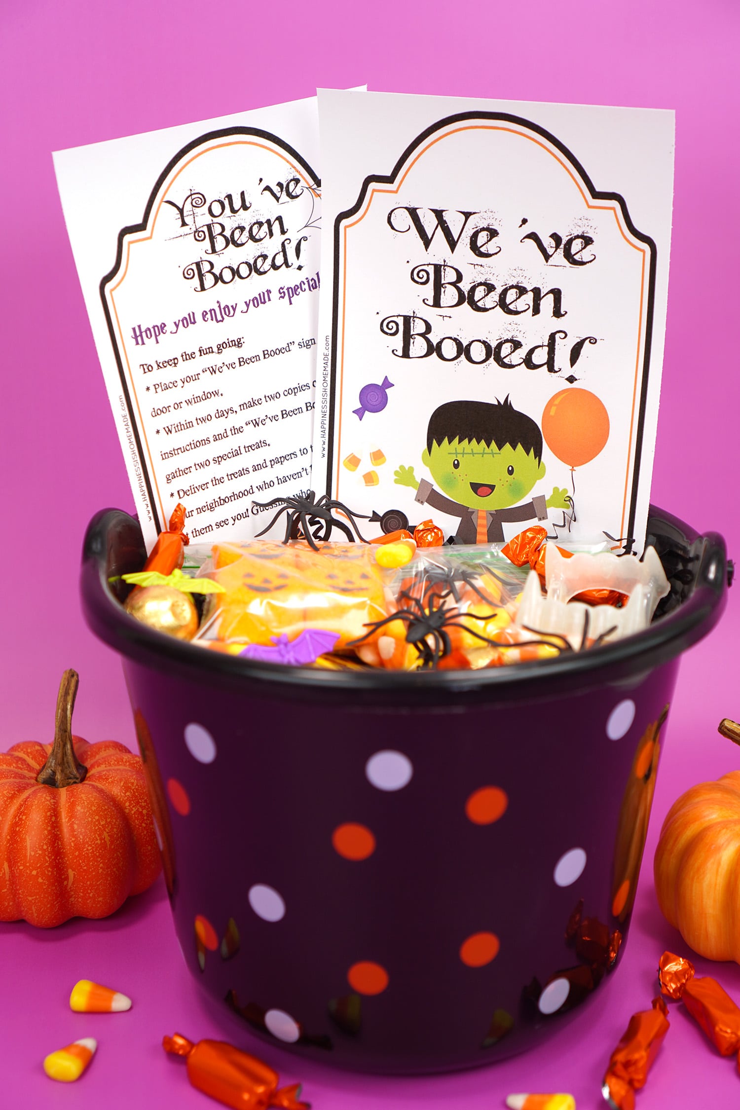 Black plastic Boo Basket filled with Hallowen candy and novelty toys with "You've Been Booed" and " We've Been Booed" printable signs on purple background with pumpkins and candy corn