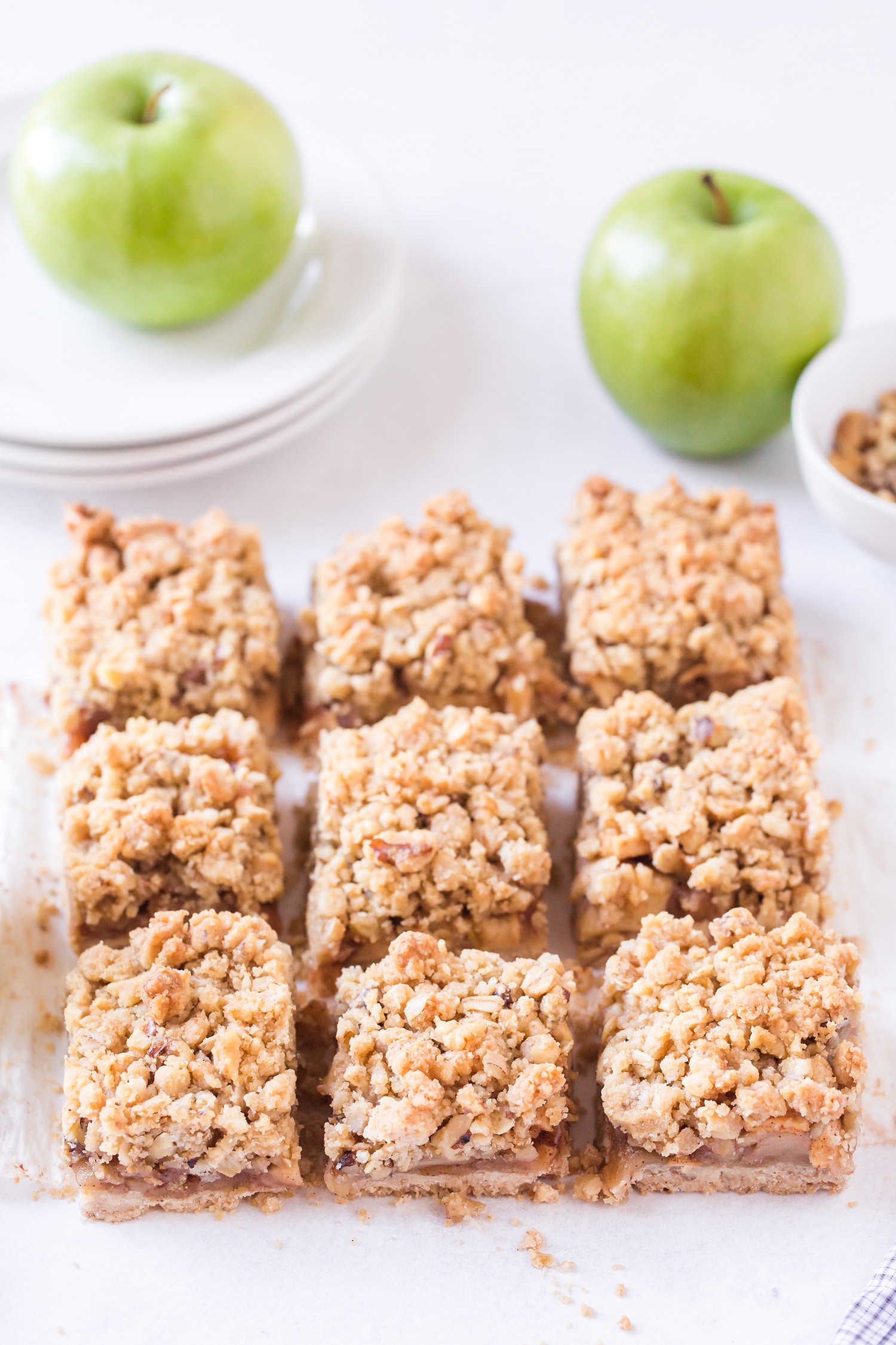 9 sliced apple pie bars on white background with apples and plates in background