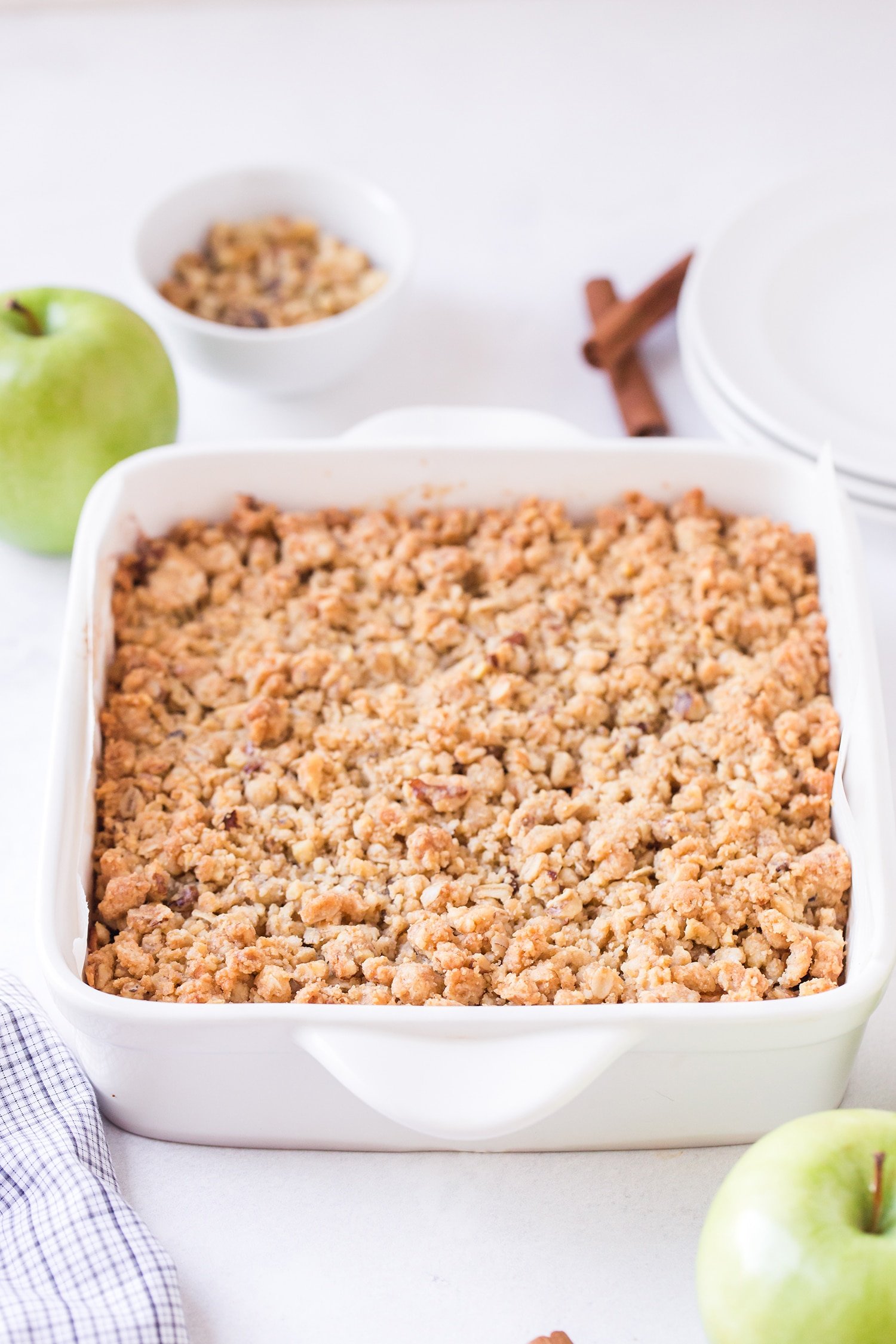 Yummy Apple Pie Bars in a white baking dish with apples and cinnamon sticks in background