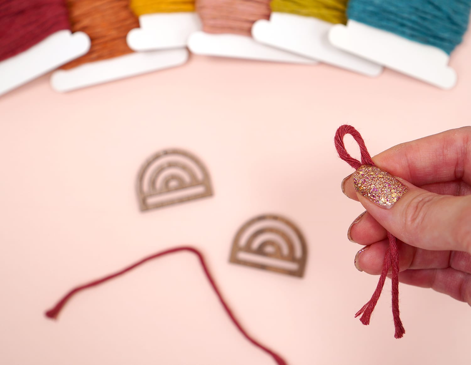 Hand holding loop of dark red macrame cord with wooden rainbow and cording skeins in background