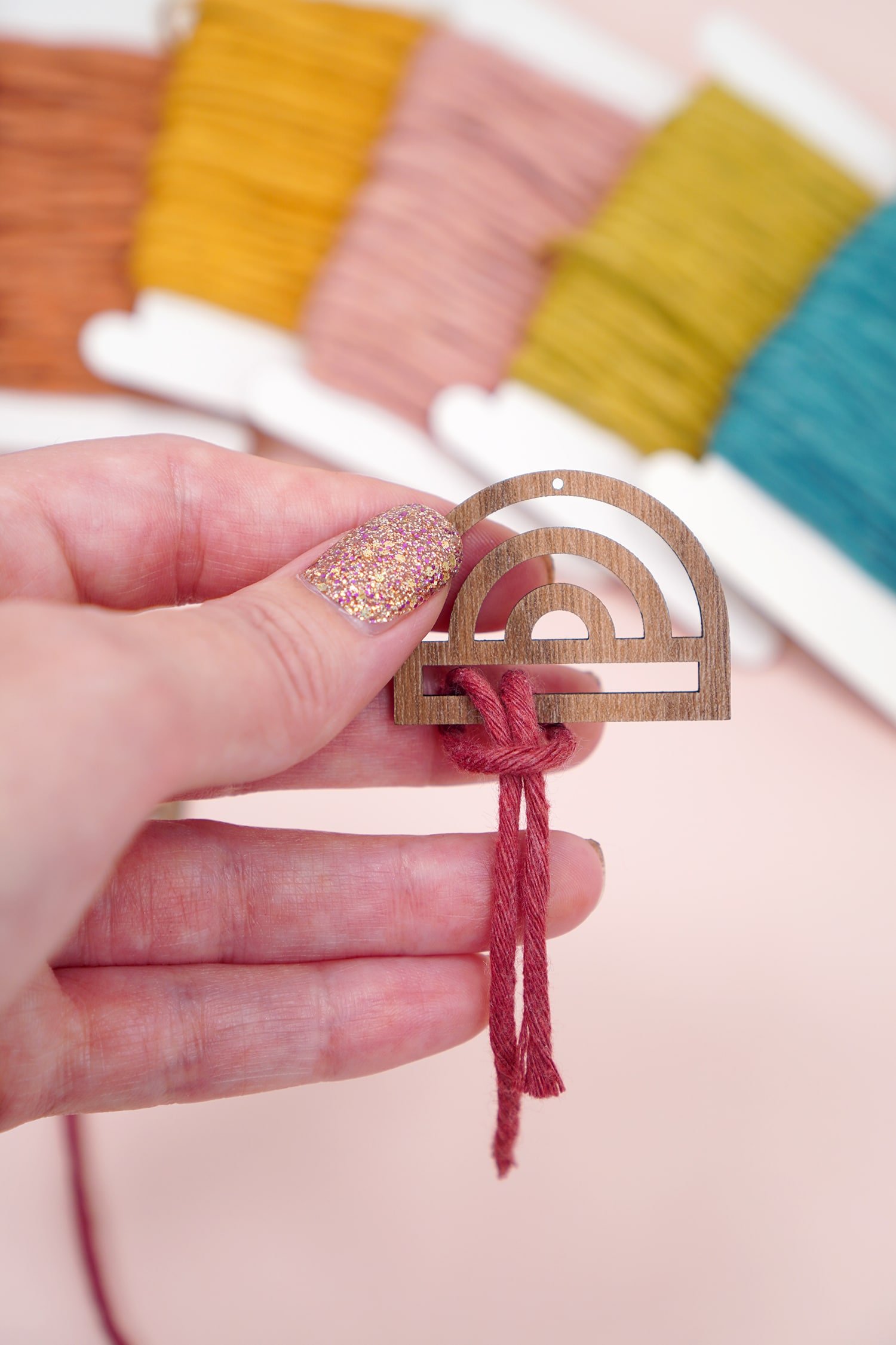 Hand attaching macrame cord to a wooden rainbow earring with macrame cord in background