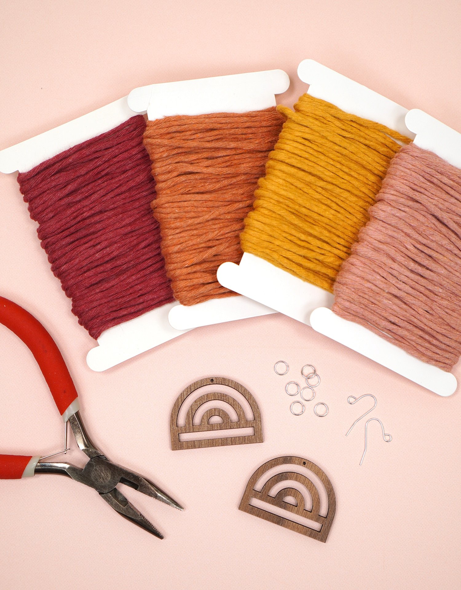 Supplies to Make Macrame Rainbow Earrings on peach background - cord, wooden rainbows, earring findings, and jewelry pliers