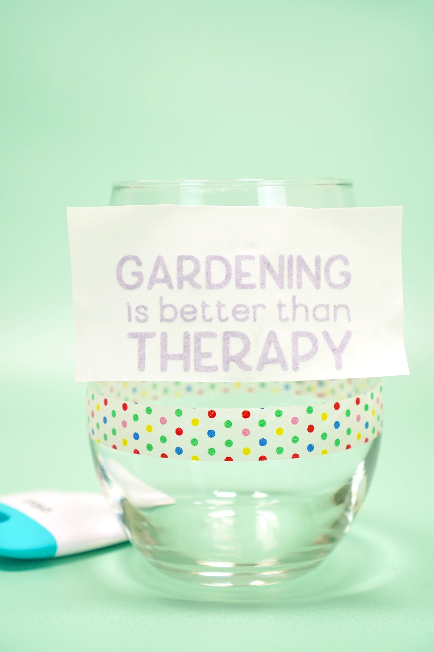 Applying "Gardening is Better Than Therapy" purple shimmer vinyl to a clear stemless wine glass on mint green background
