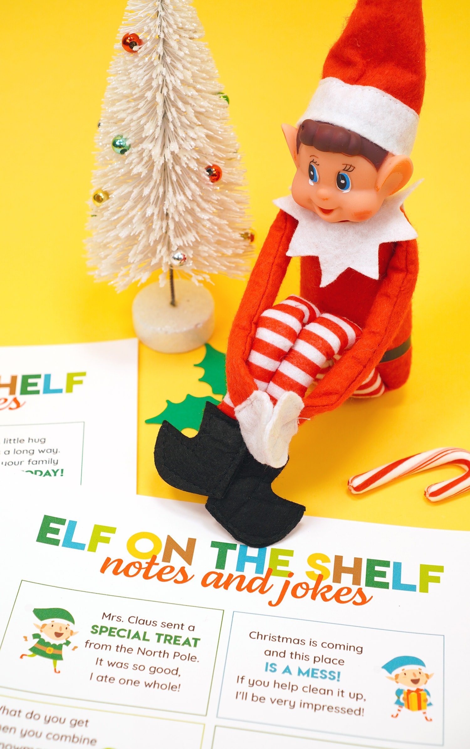 Elf doll on a yellow background with printed Elf on the Shelf note and joke cards and candy canes and Christmas decorations in the background.