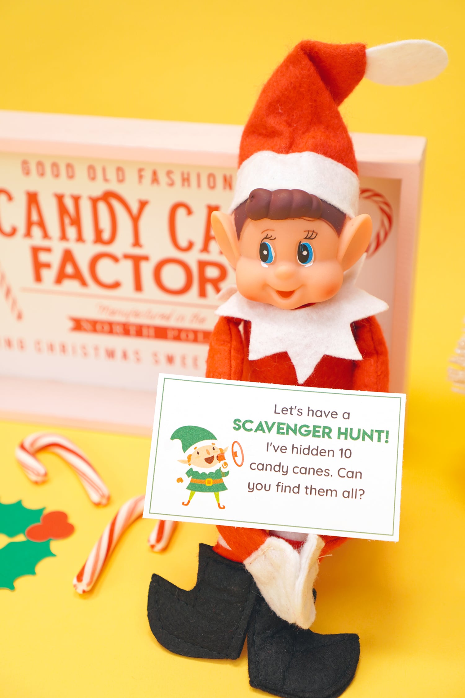 Elf doll on a yellow background holding a printed Elf on the Shelf scavenger hunt note card and candy canes and Christmas decorations in the background.