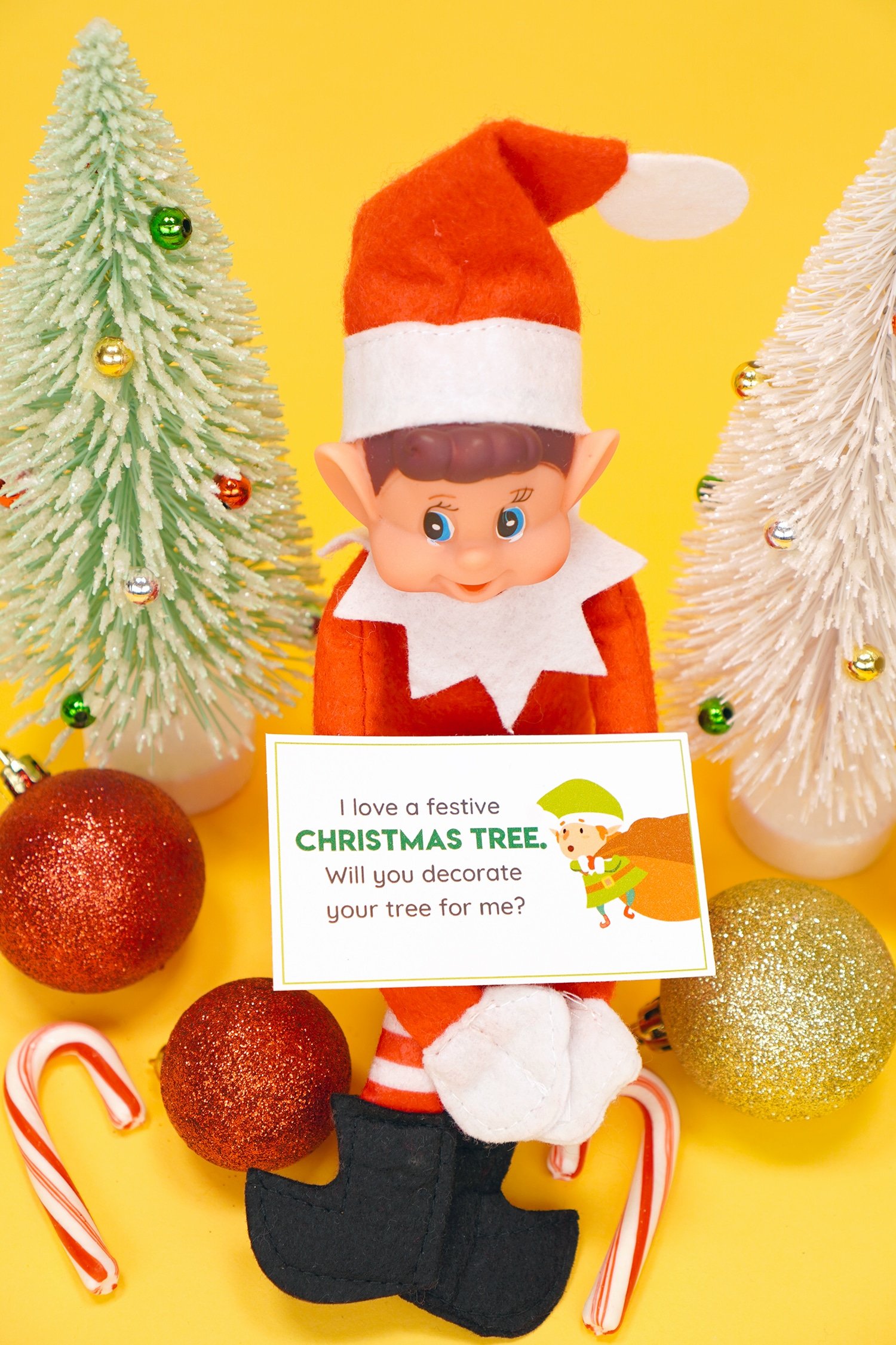 Elf doll holding a printable Elf on the Shelf note card with candy canes, ornaments, and glitter Christmas trees on yellow background