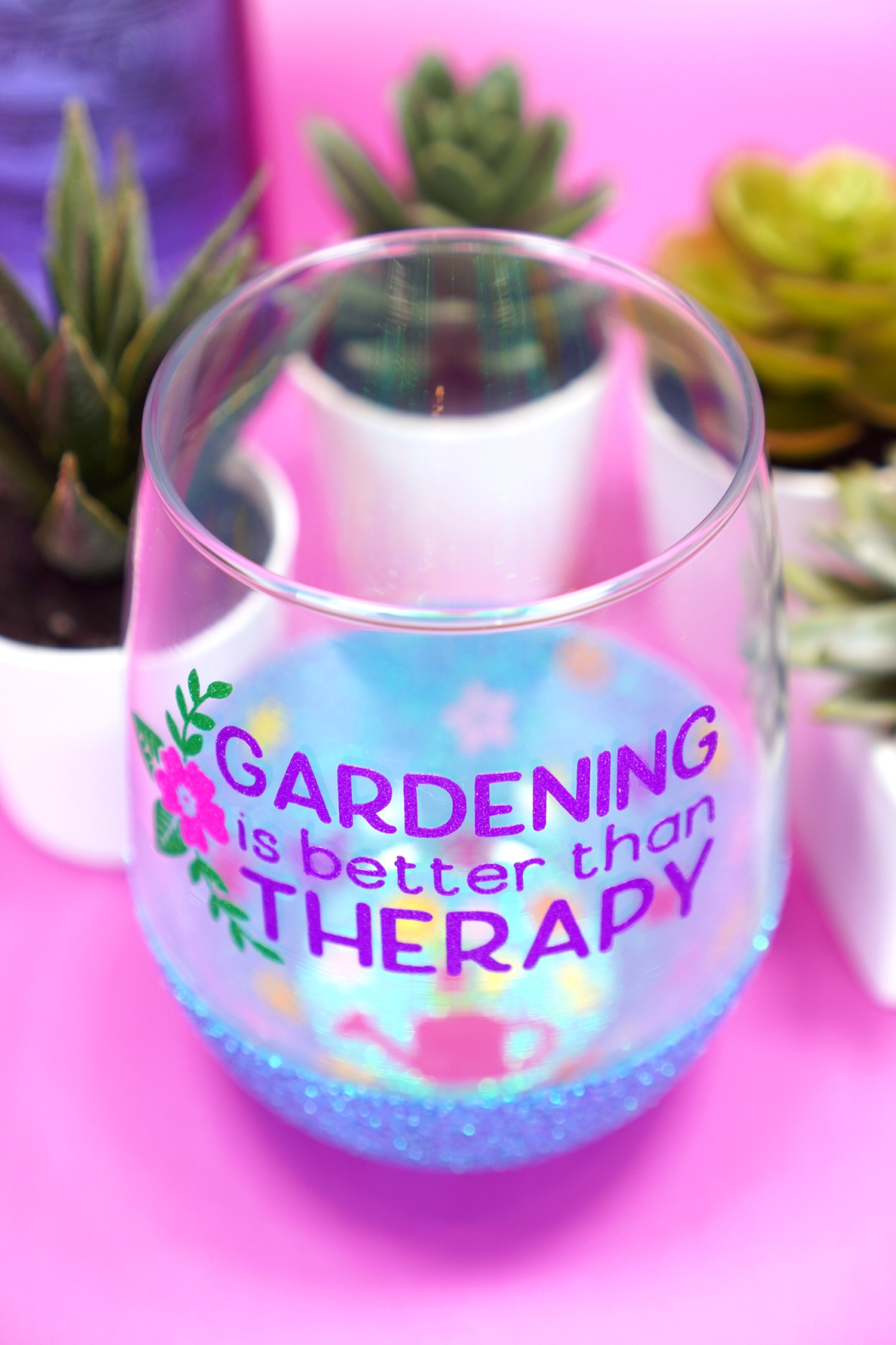 Overhead angle shot of "Gardening is Better Than Therapy" wine glass with blue glittered base and a peekaboo watering can and flowers design inside