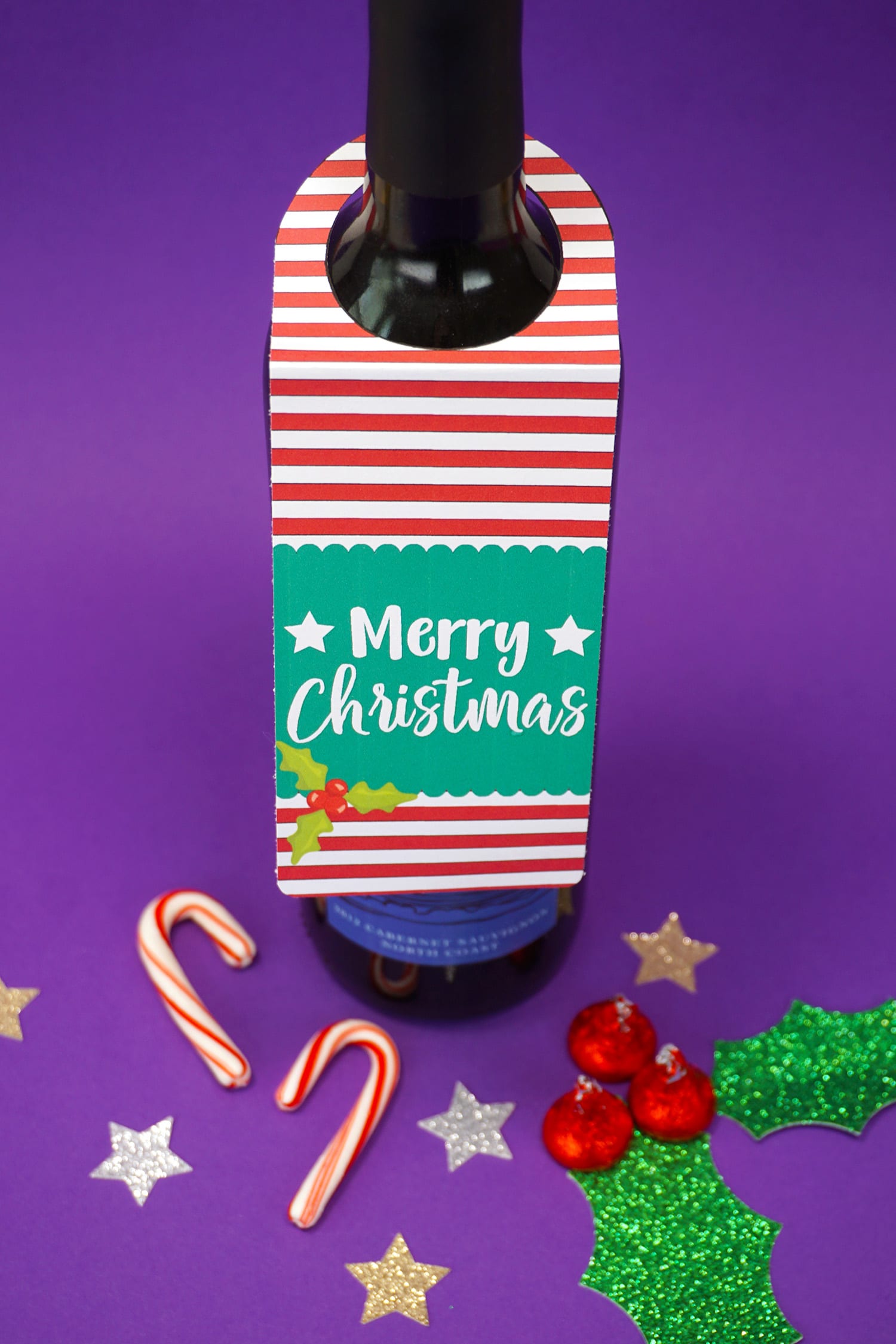 "Merry Christmas" red, white, and green Christmas wine tag on bottle on purple background with candy canes and stars