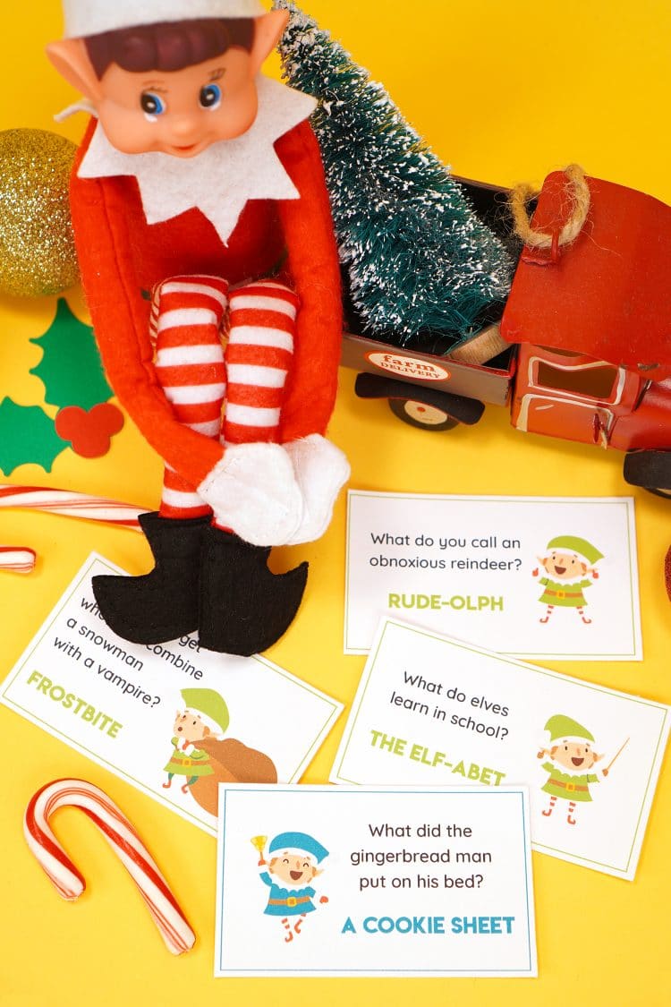 Elf doll on a yellow background with printed Elf on the Shelf joke cards scattered around him and candy canes and Christmas decorations in the background. 