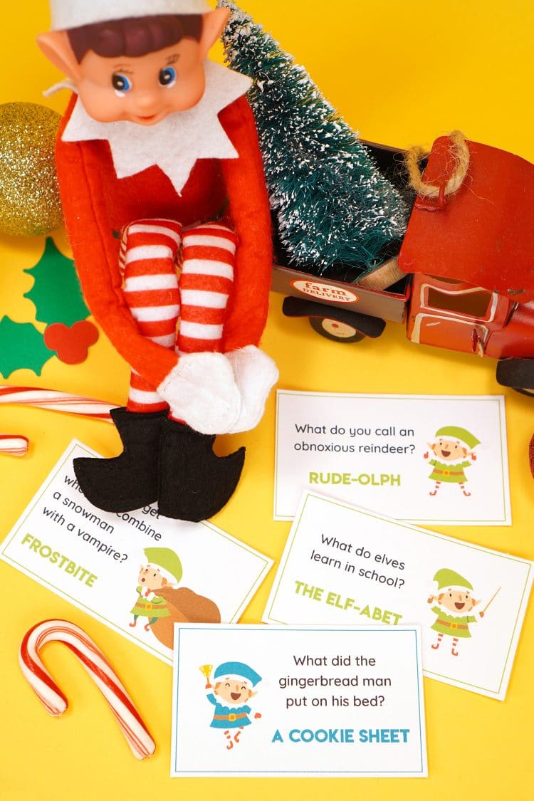 Elf doll on a yellow background with printed Elf on the Shelf joke cards scattered around him and candy canes and Christmas decorations in the background.