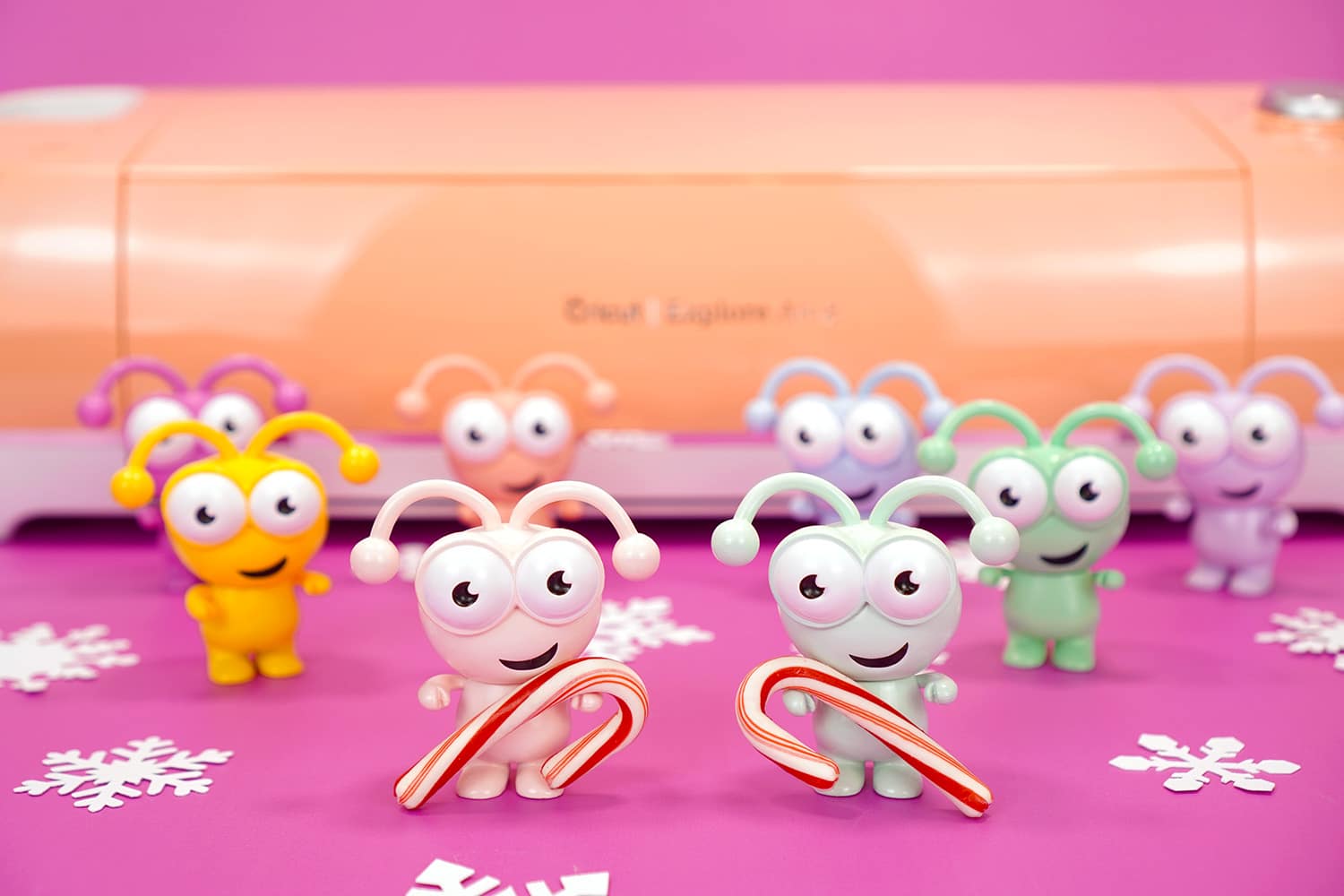 Rainbow colored Cricut Cutie figures holding candy canes on purple background with peach Cricut Explore Air 2 machine