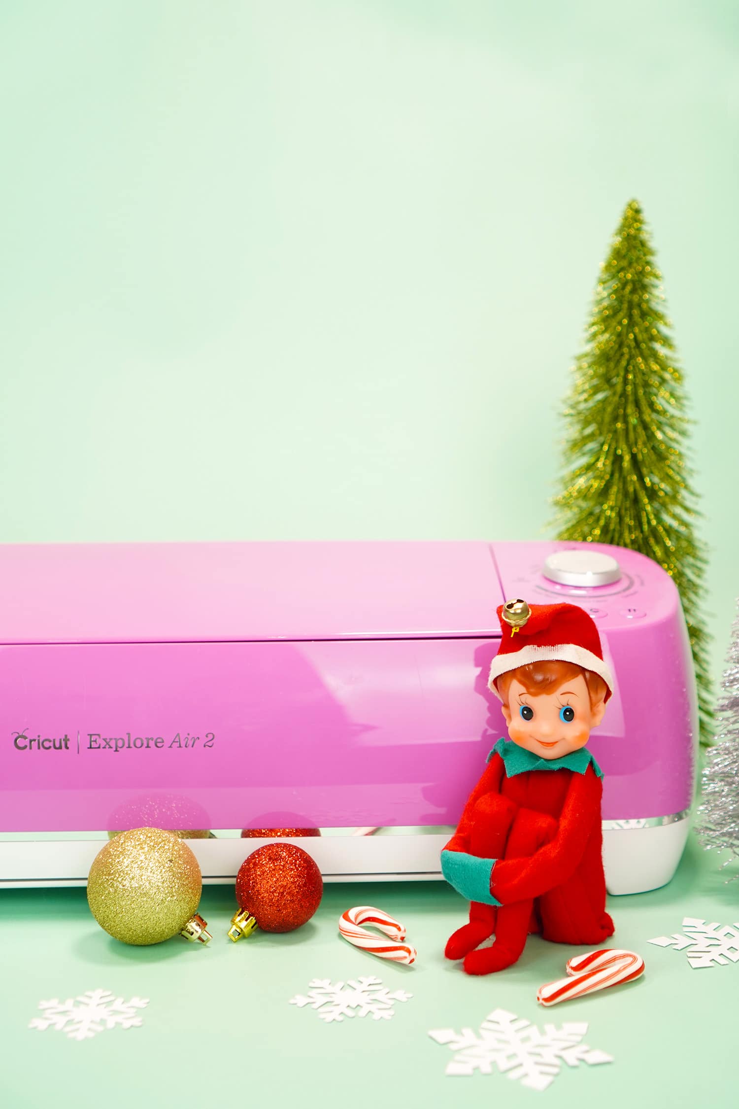 Vintage Elf in front of pink Cricut Explore Air machine on mint background with ornaments and bottle brush trees