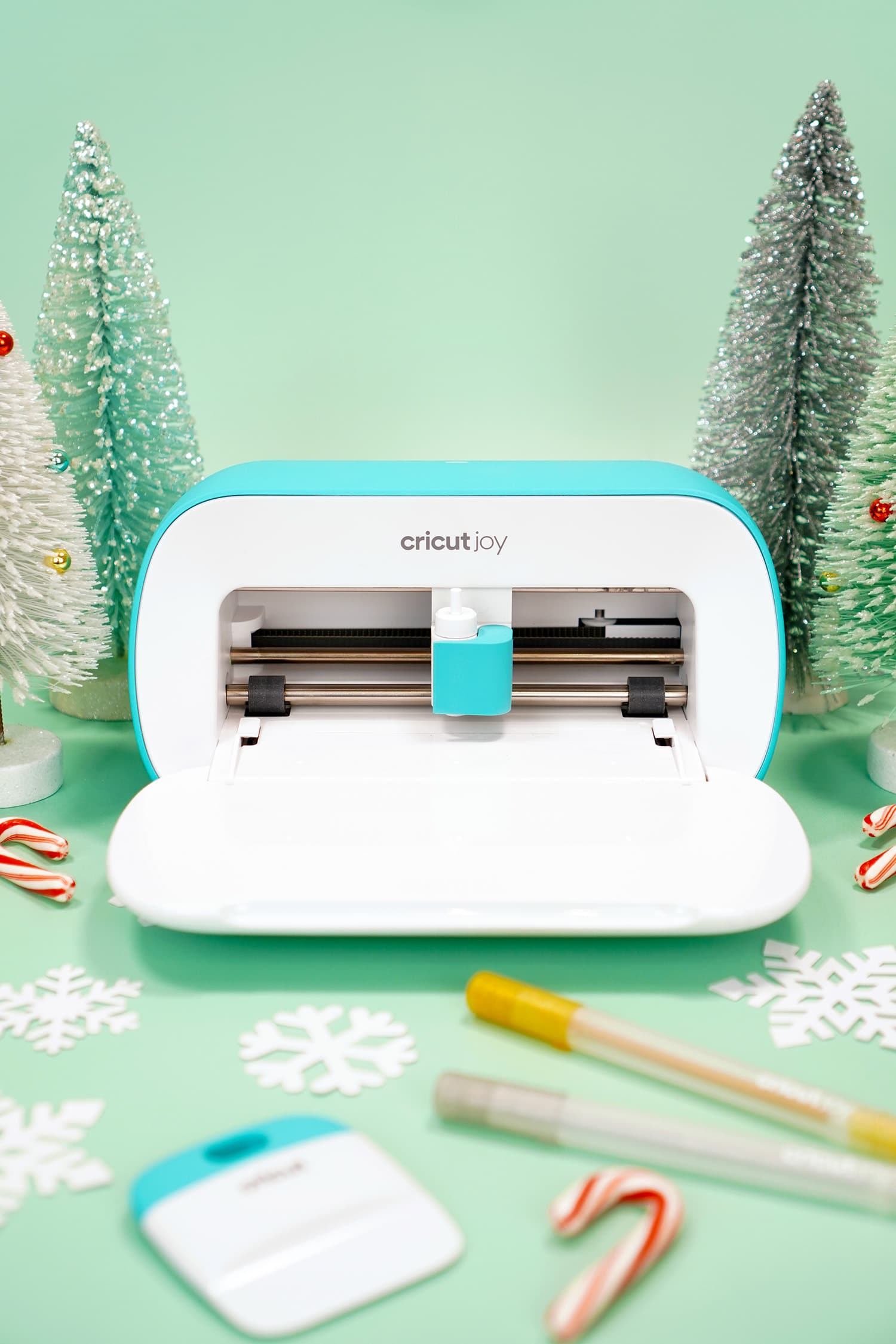 Cricut Joy machine surrounded by Cricut tools, paper snowflakes, and candy canes on mint green background