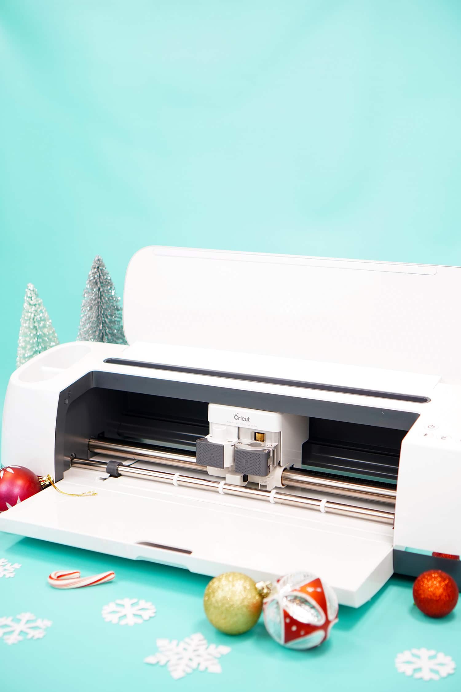 Cricut Maker machine on aqua background with white snowflakes and red and gold ornaments