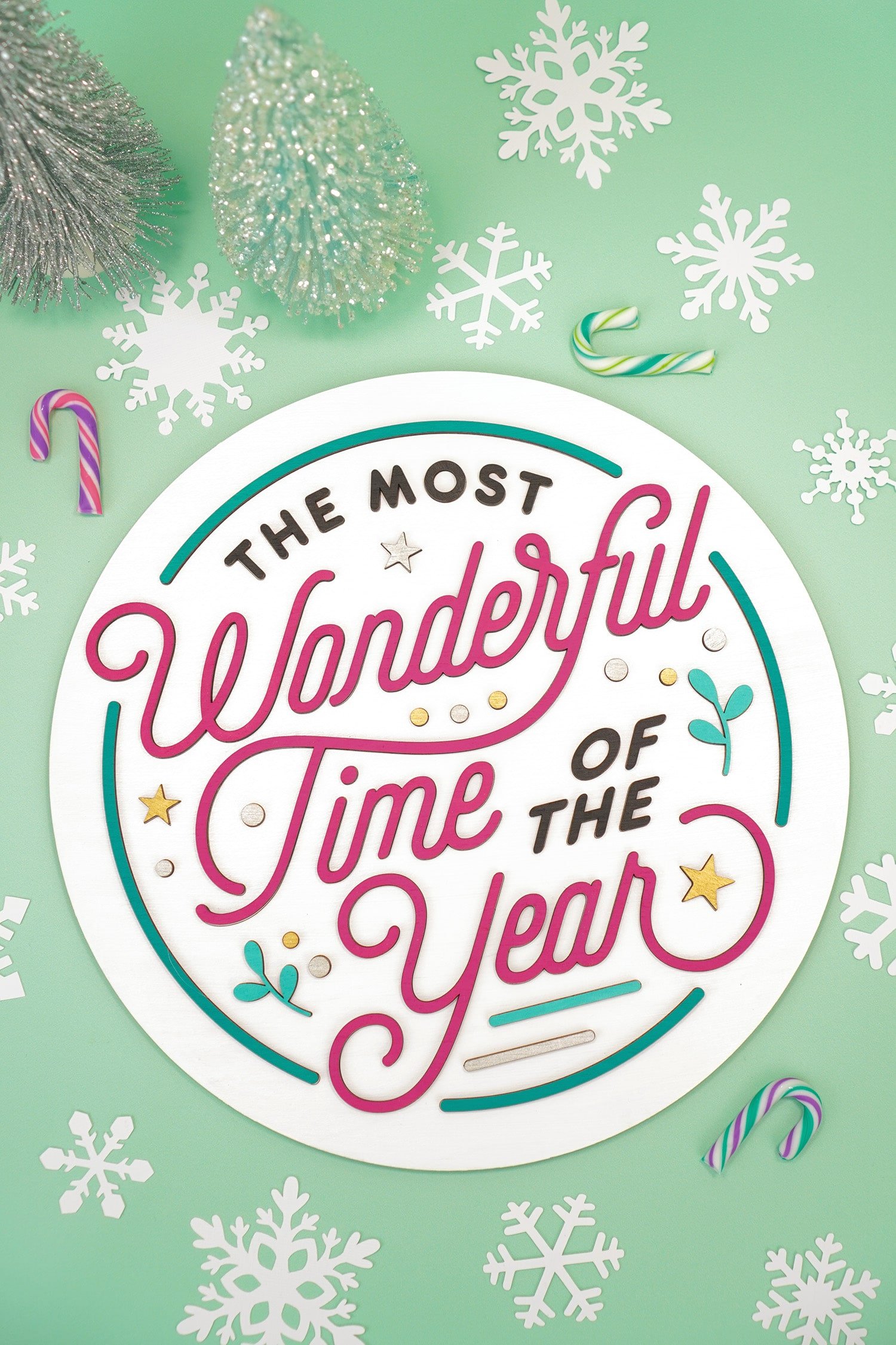 Round layered 3D laser cut wood sign \"The Most Wonderful Time of the Year\" on mint green background with colorful candy canes, white paper snowflakes