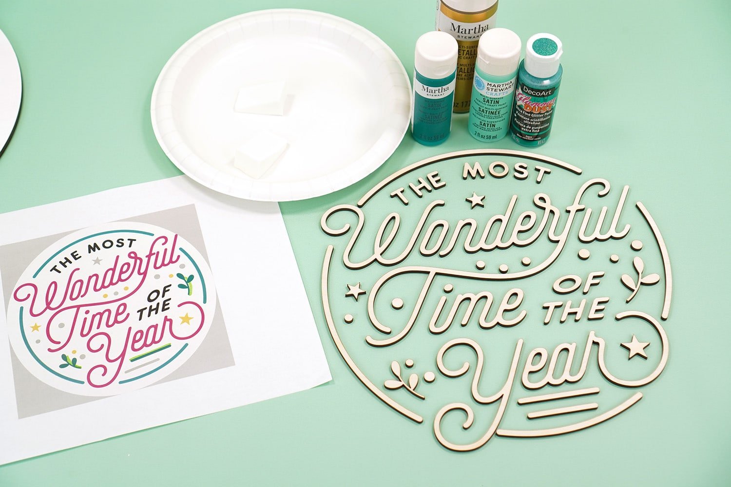 Mint green background with "The Most Wonderful Time of the Year" Glowforge laser cut wood pieces, paint, and a print out of color palette on white paper